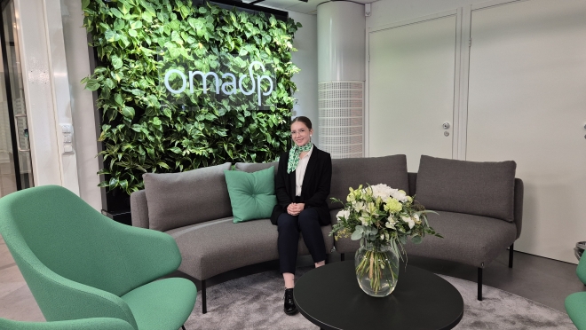 Kuvassa sohvalla Paula Hautamäki, taustalla viherseinä, jossa OmaSp:n logo valkoisella.