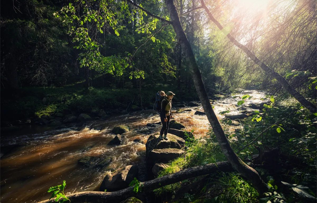 Nainen lapsi selässään kesäisessä puromaisemassa.